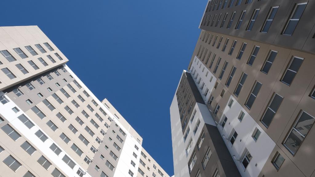 Imagem de dois edifícios residenicias tirada de baixo pra cima com um ceu azul ao fundo