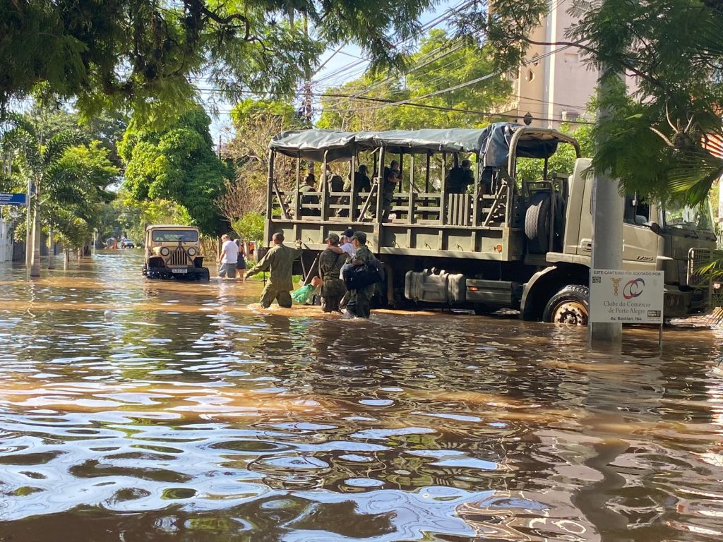 Rua alagada com caminhões para o resgate de pessoas. Agentes e voluntários se moviementam na água para ajudar pessoas isoladas.