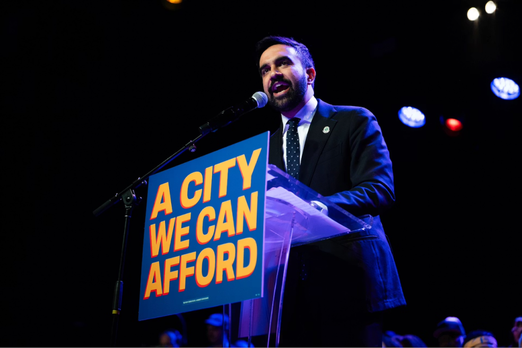 O candidato à prefeitura de Nova York, Zohran Mamdani dircusando em evento de campanha. No púlpito onde faz sua fala tem um cartaz com a frase em inglês: "A CITY WE CAN AFFORD", toda em caixa alta.
